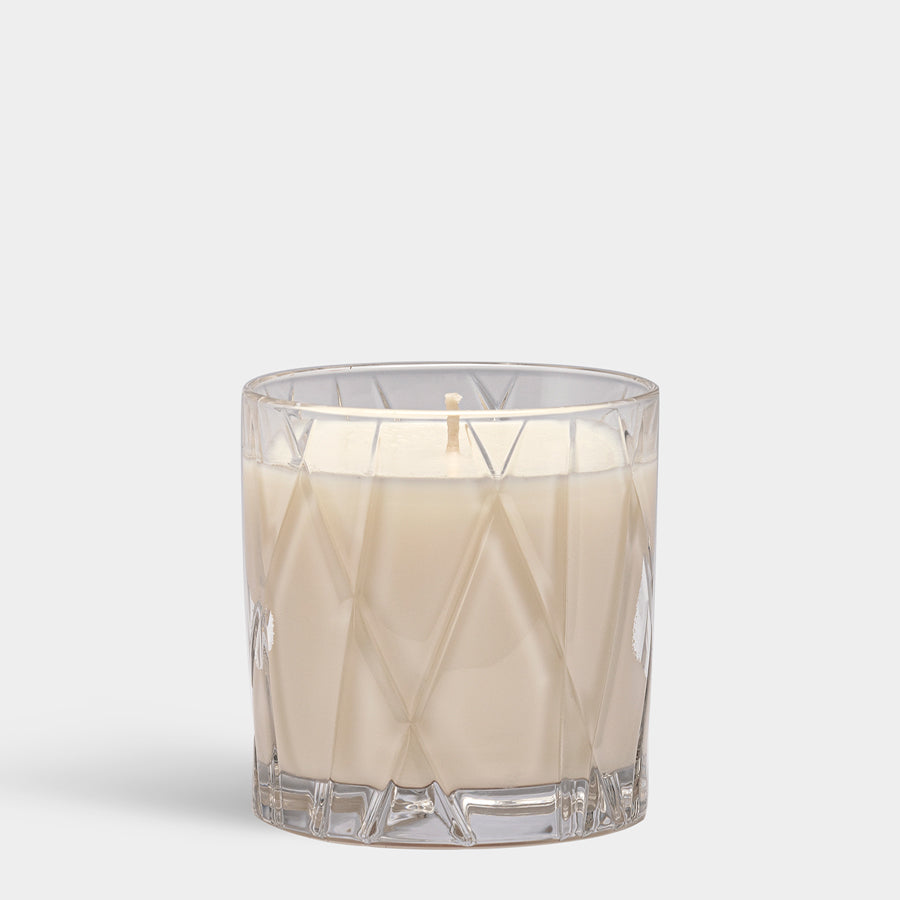 A white Orrefors: City Candle - Warm Amber and Oakmoss, in a textured glass holder, stands elegantly against a plain gray background.