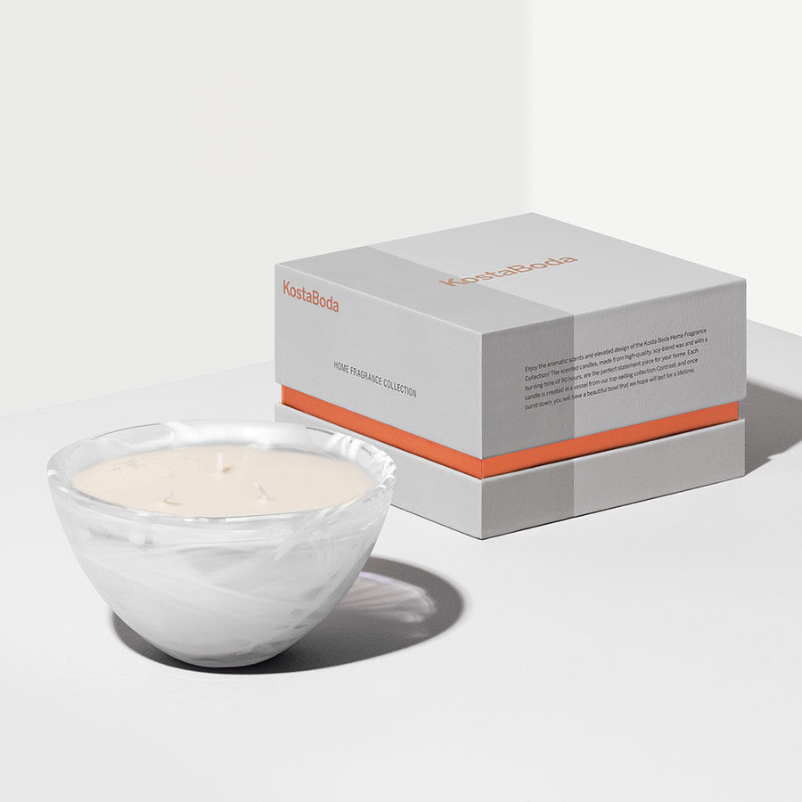 A KostaBoda Home Fragrance Collection Contrast White Candle in Woodland Vanilla scent rests in a white marble-patterned bowl on a white surface, paired with its gray and orange box, adding serene elegance to the scene.