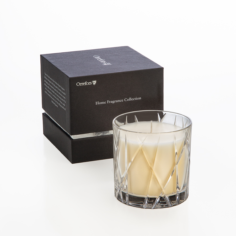 An Orrefors City Candle in a faceted glass holder sits elegantly beside a black box labeled Home Fragrance Collection, infused with rich warm amber and oakmoss notes.