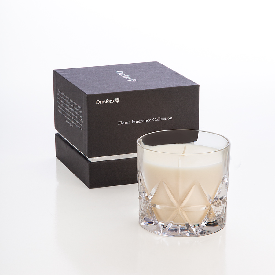 An Orrefors Peak Candle in Woodland Vanilla rests before a black Home Fragrance Collection box, releasing its soothing aroma.