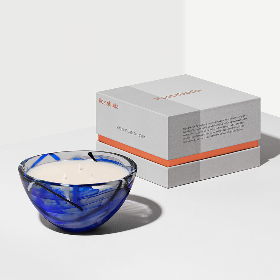 The KostaBoda: Contrast Candle Blue, in a blue glass bowl beside its labeled white box, adds a coastal bloom to your home fragrance collection.