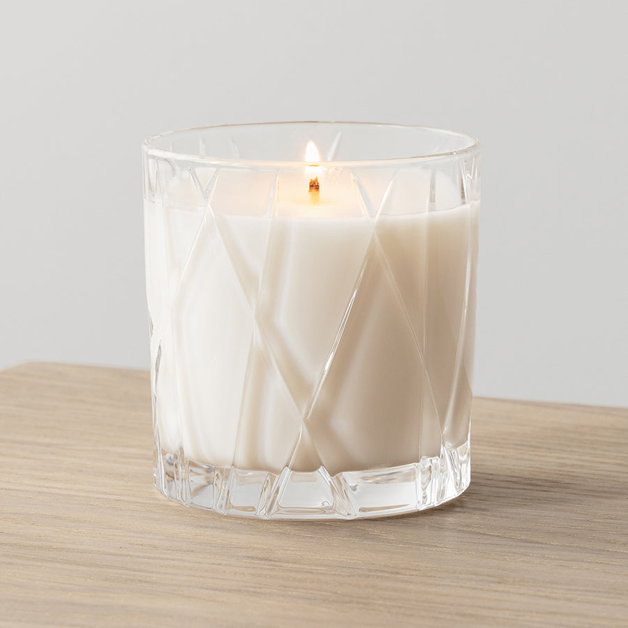 An Orrefors City Candle in a clear faceted glass holder sits on wood, releasing warm amber and oakmoss scents.