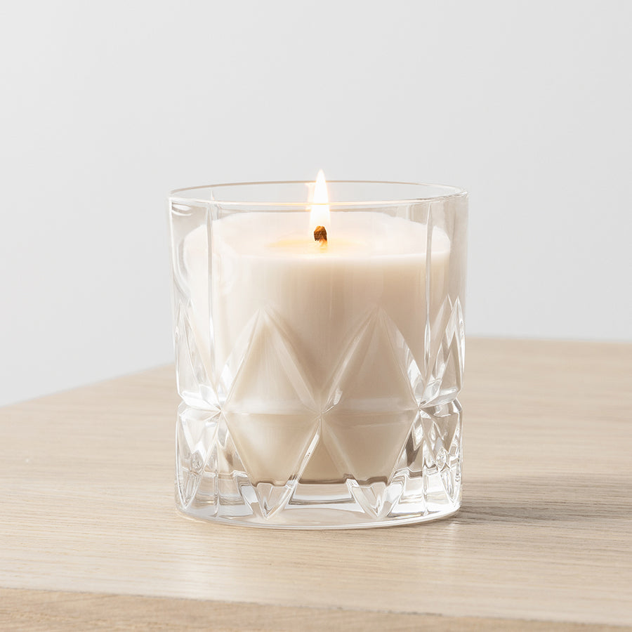 The Orrefors: Home Fragrance Collection Peak Candle in Woodland Vanilla, set in a faceted glass container, sits on wood, adding coziness to your collection against a plain background.