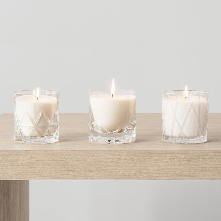 Three Orrefors: City Candles - Warm Amber and Oakmoss, sit in glass holders on a light wood surface, emitting a subtle fragrance against a plain backdrop.