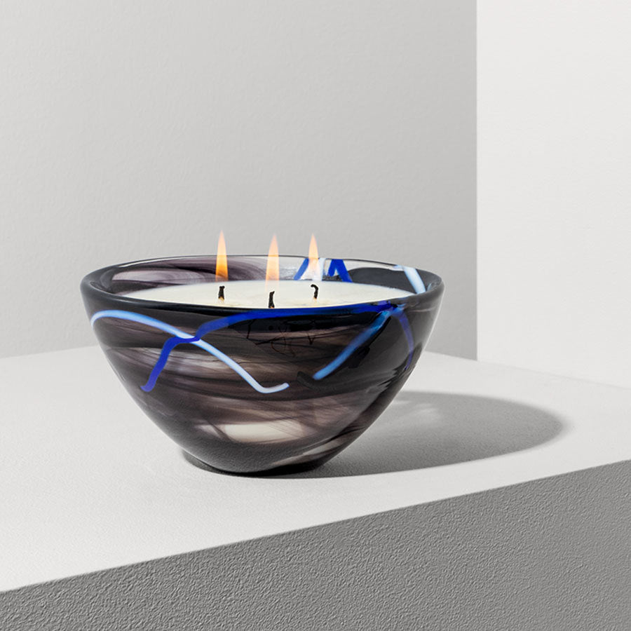 The KostaBoda Contrast Black Candle from the Home Fragrance Collection showcases a swirled black and blushing pink design, cradling three lit wicks. It elegantly rests on a light surface, enhancing its rich hues.