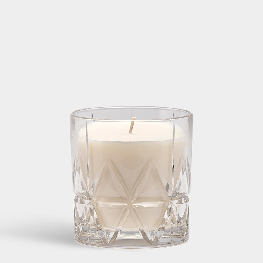 A Woodland Vanilla candle from the Orrefors: Home Fragrance Collection Peak sits in a clear, patterned glass holder against a plain gray background.