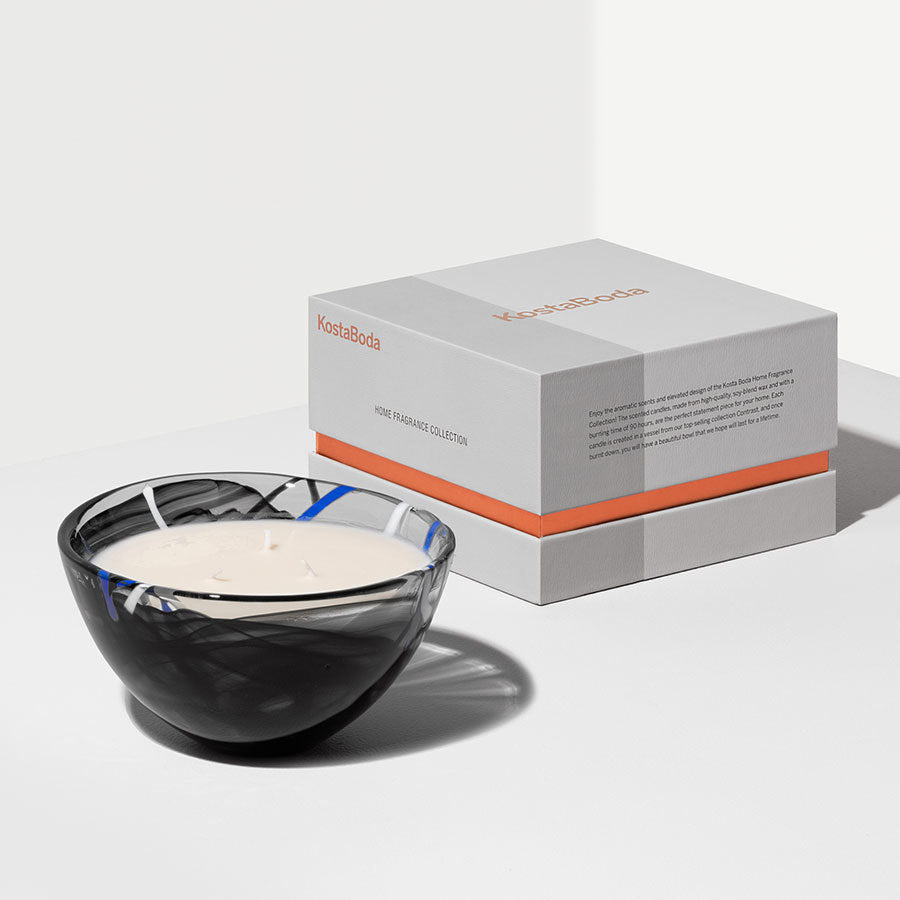 A KostaBoda Contrast Black Candle from the Home Fragrance Collection, featuring a black and white glass bowl with two wicks in Blushing Pink, sits beside a closed gray box with orange accents.