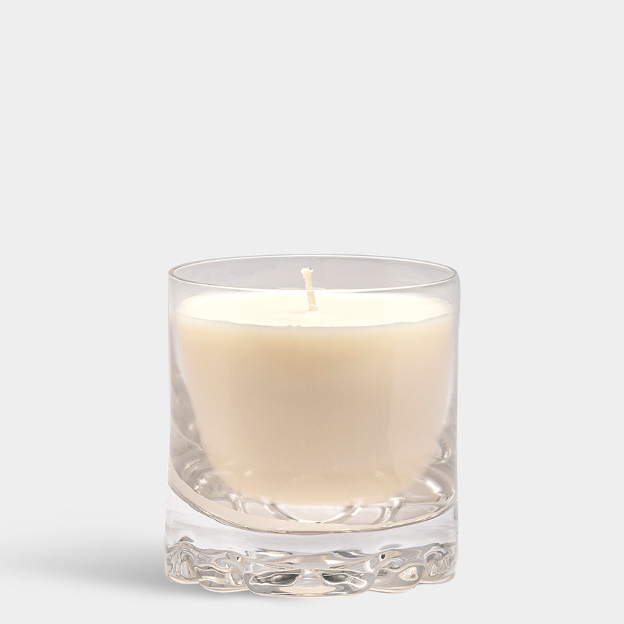 The Orrefors: Erik Candle Blushing Pink, featuring a subtle floral scent, is elegantly presented in a white hue nestled within a clear Orrefors glass holder against a plain background.