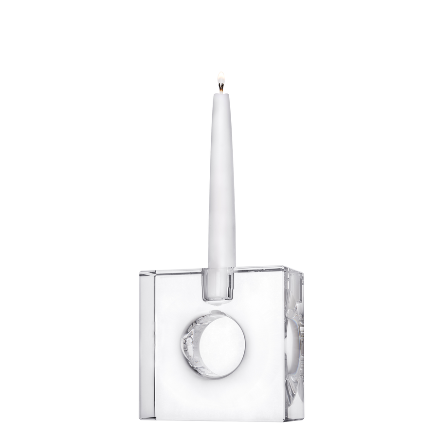 A tall, lit candle in the Orrefors: Torg Square Candle Holder by Karim Rashid, showcasing a translucent decorative finish.