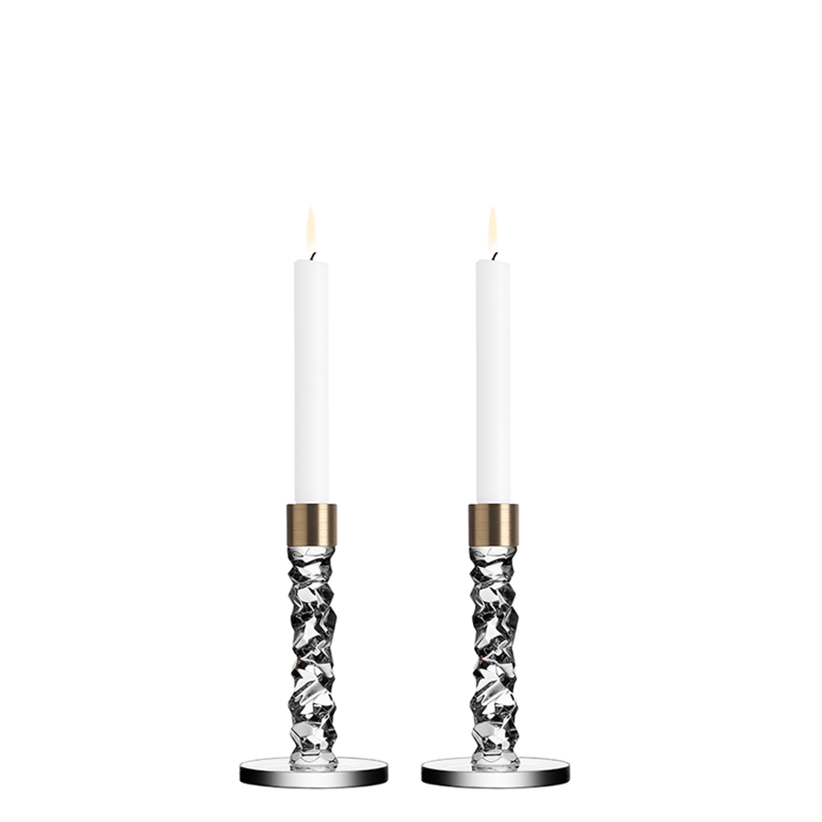 Two lit candles are set in Orrefors Carat Candlestick Brass holders, their textured glass and brass accents exuding elegance against a plain background.