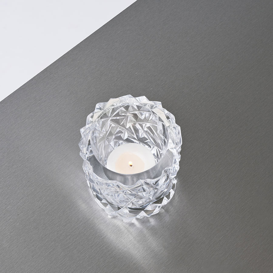 A small white candle elegantly rests in an Orrefors: Carat Candle Holder Medium, placed on a smooth gray surface.