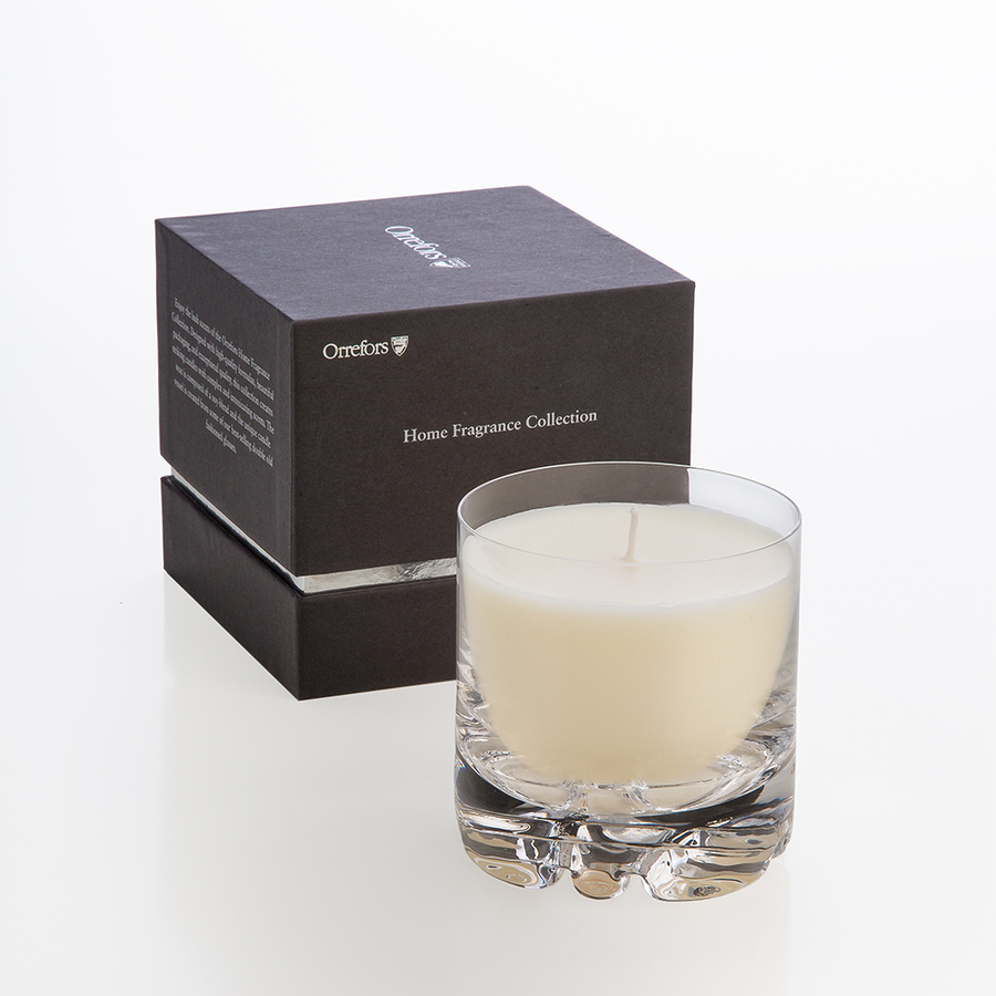 The Orrefors: Erik Candle Blushing Pink rests elegantly in front of a sleek black box labeled Home Fragrance Collection, releasing a delicate floral aroma into the air.