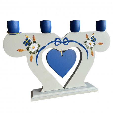 A handcrafted wooden candle holder from Sweden, featuring a white design with blue candle holders, adorned with Swedish flag motifs and floral patterns, includes a central blue heart.