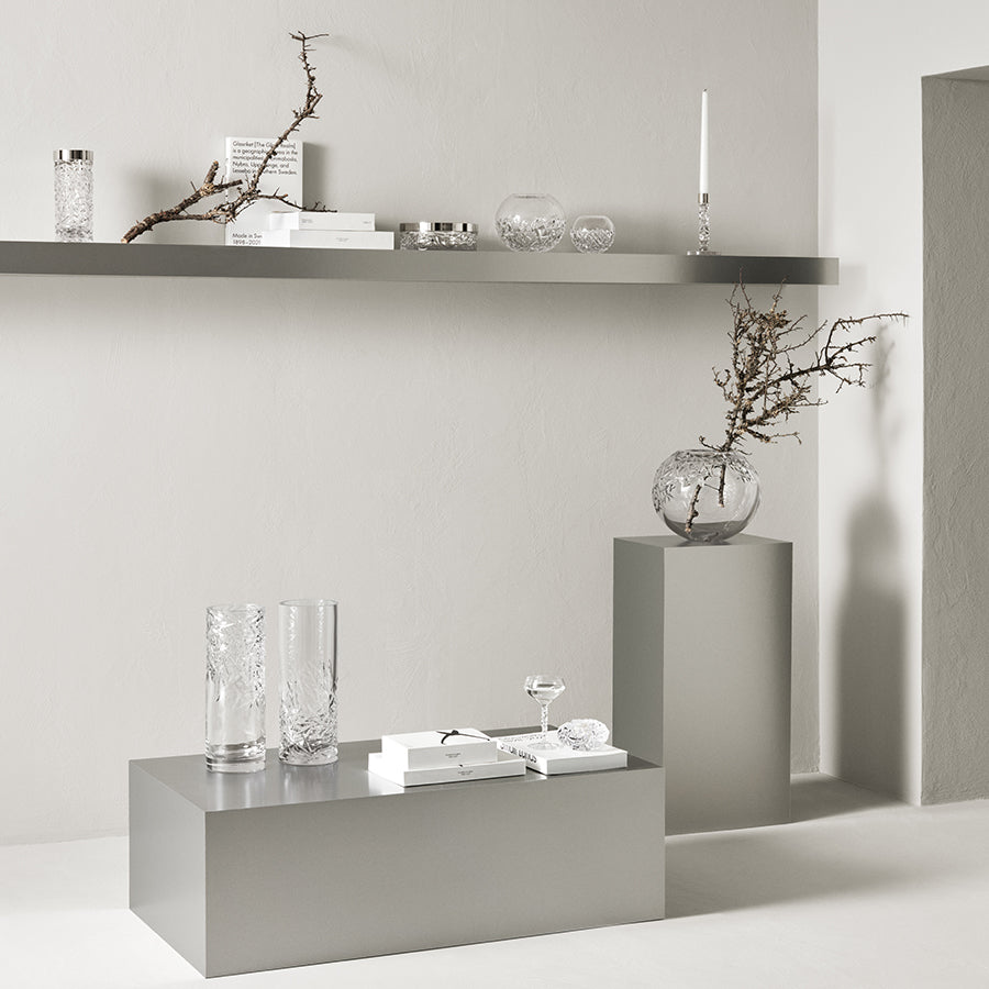 The minimalist interior includes two pedestals and a shelf with clear crystal glass vases, books, and an Orrefors: Carat Candlestick Silver Small. Sparse dried branches in vases add a natural touch, with muted tones emphasizing the elegance of the Carat collection by Orrefors.