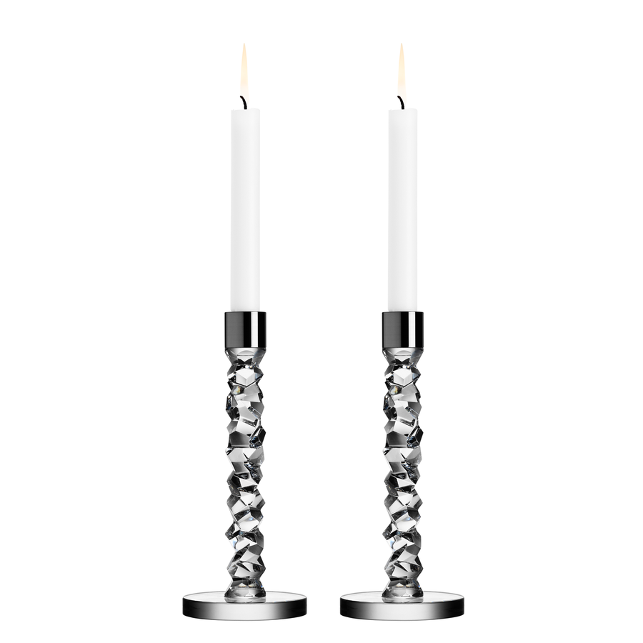 The Orrefors Carat Candlestick Silver Medium (2-Pack) features two lit, white candles in sophisticated, crystal glass geometric holders with silver bases, evoking a design reminiscent of Lena Bergströms artistry.