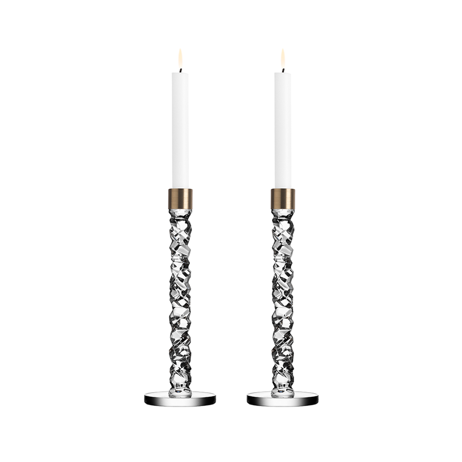 Two tall, lit Orrefors: Carat Candlestick Brass Large candles in intricately designed silver holders with brass accents set against a white background.