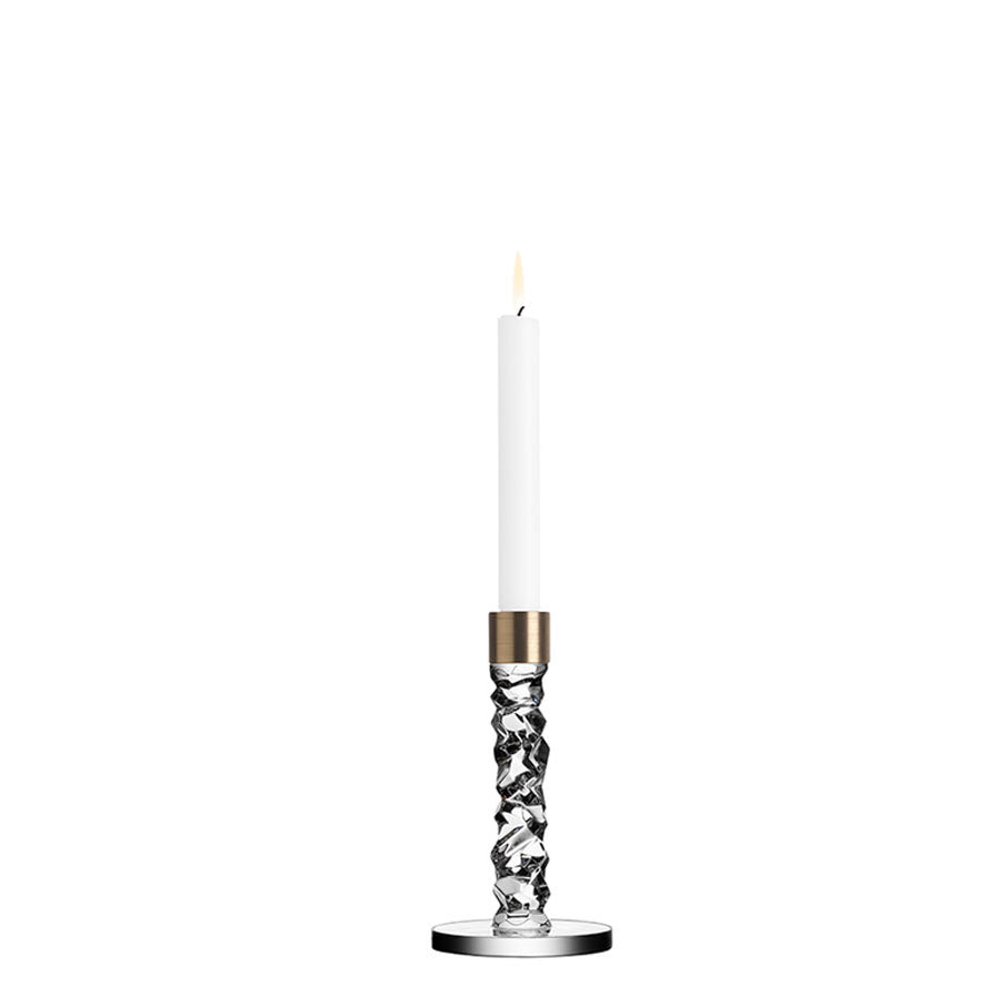 A lit white candle elegantly rests on the Orrefors Carat Candlestick Brass Small, featuring a metallic accent.