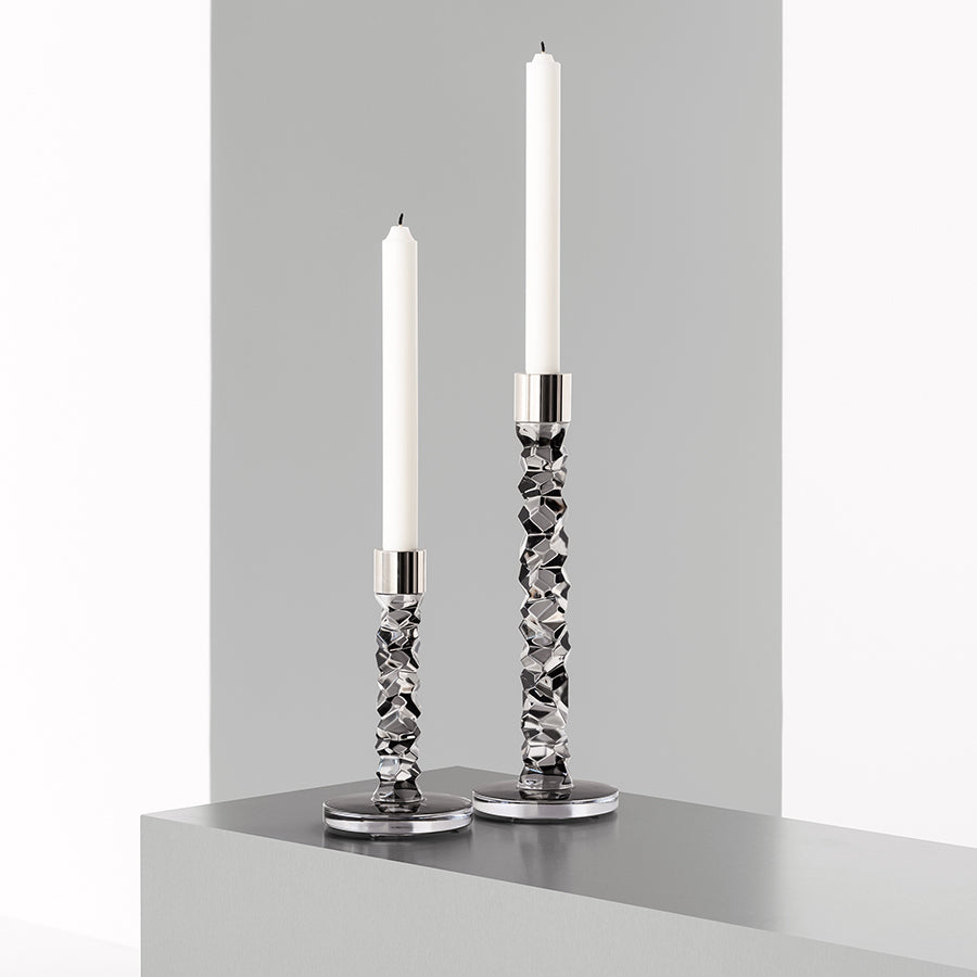 Two large Orrefors Carat Graphite candlesticks rest on a gray surface, showcasing textured bases. Each holds a tall white candle, casting a serene glow. The plain, light background enhances the refined simplicity Lena Bergström might envision.