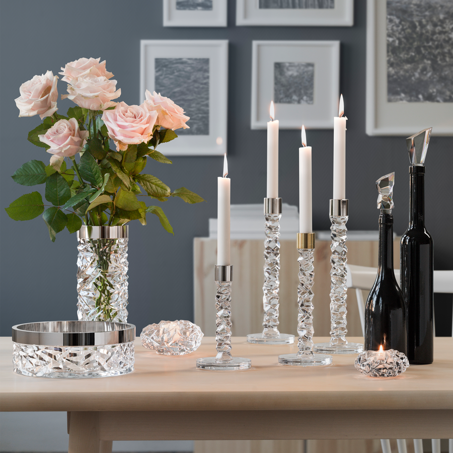 Elegant table setting with pink roses in a vase, Orrefors Carat Candlestick Brass Medium (2-Pack) holding lit white candles, a crystal bowl, and black wine bottles against a backdrop of framed art.