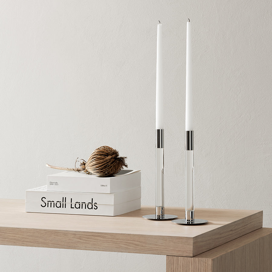 A pair of Orrefors Lumiere Candlestick Silver Medium holders with tall white candles sits on a wooden table adorned with books and a decorative straw piece, against a neutral wall.