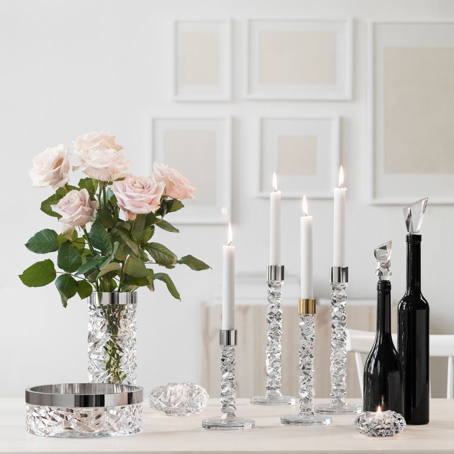 A table adorned with pink roses in an Orrefors crystal vase is enhanced by five lit Orrefors Carat Candlestick Silver Small, alongside black bottles. Nearby, a crystal dish sparkles, while white framed art elegantly hangs against the white wall.