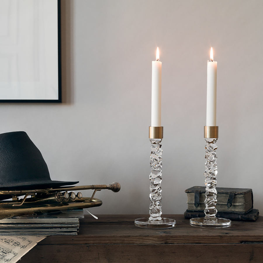 Two Orrefors Carat Candlestick Brass Large holders rest elegantly with a sleek black hat, gleaming brass trumpet, and stacked books against a muted gray wall.