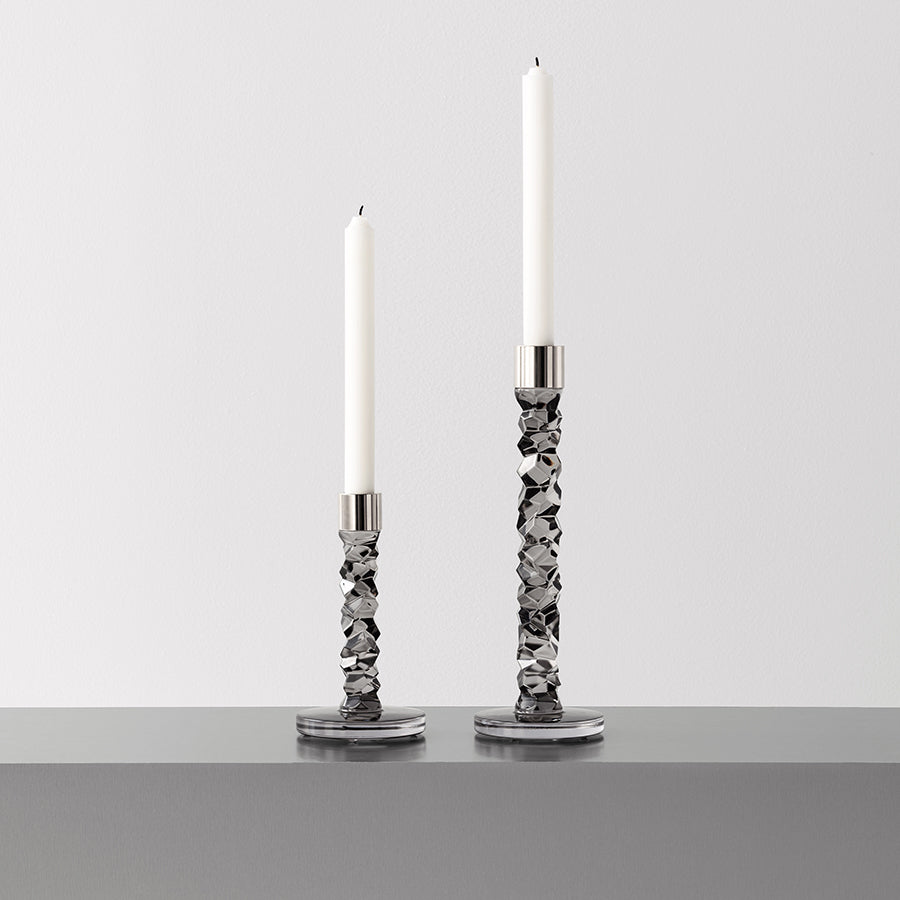 Two Orrefors Carat Graphite Candlestick Large holders with tall white candles grace a gray surface. Designed by Lena Bergström, they exude sophistication against the plain white background.