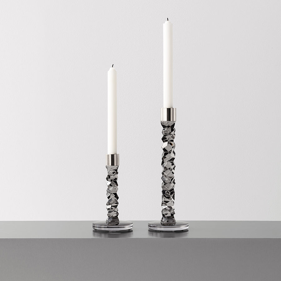 Two Orrefors Carat Graphite small candlesticks with white candles sit on a sleek gray surface against a plain white background, highlighting their elegance.