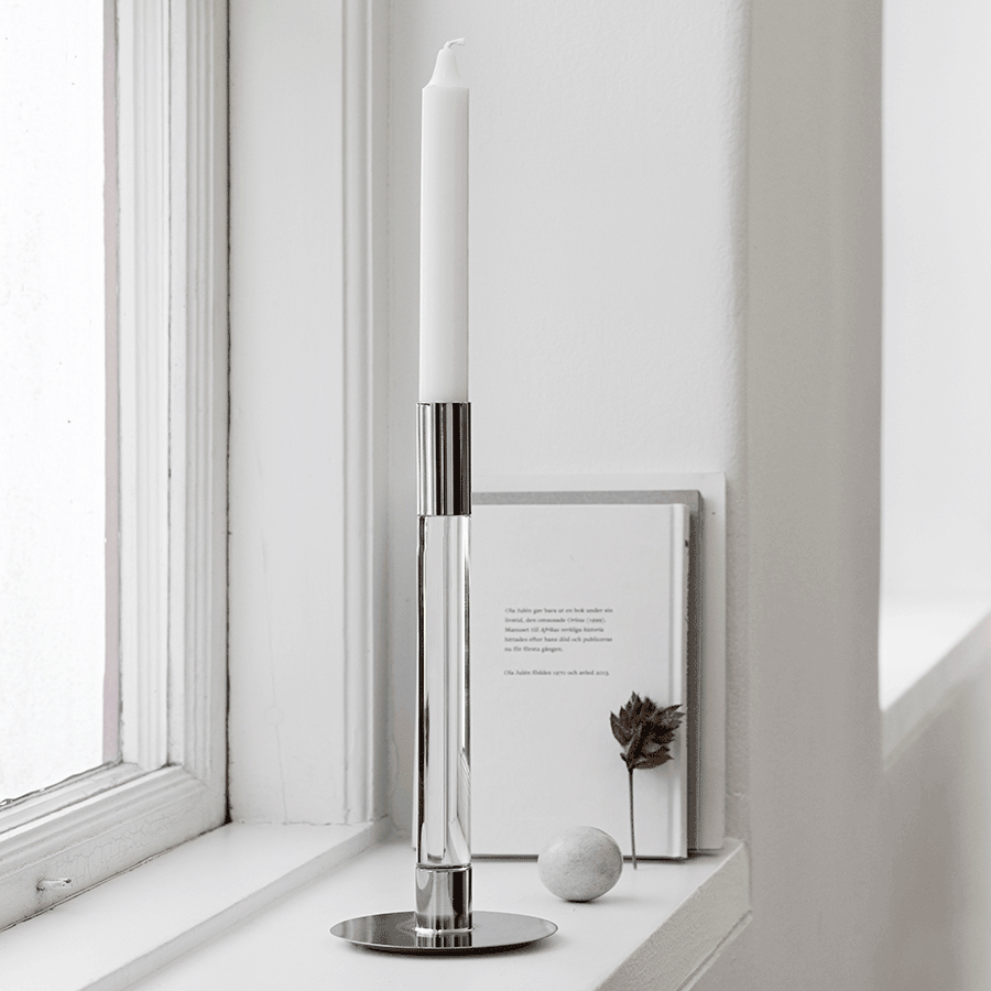 The Orrefors Lumiere Candlestick Silver Medium set, designed by Ingegerd Råman, elegantly holds a white candle on the windowsill, complementing the white book and decorative items beside it.