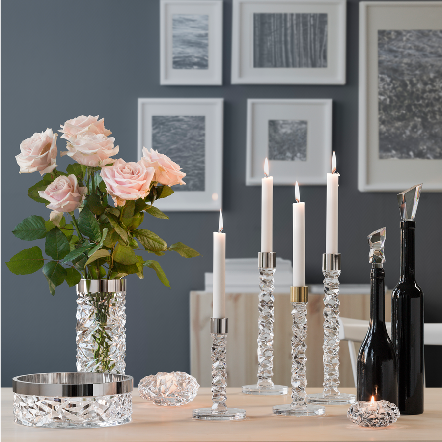 Crystal glass candle holders (Orrefors Carat Candlestick Silver Large), lit candles, a vase of pale pink roses, an Orrefors corkscrew, a wine bottle with stopper, and art frames set the scene on the wooden table.