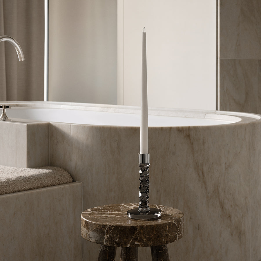 An Orrefors Carat Graphite Candlestick with a white taper candle rests on a small, round marble table beside a bathtub in a modern bathroom.