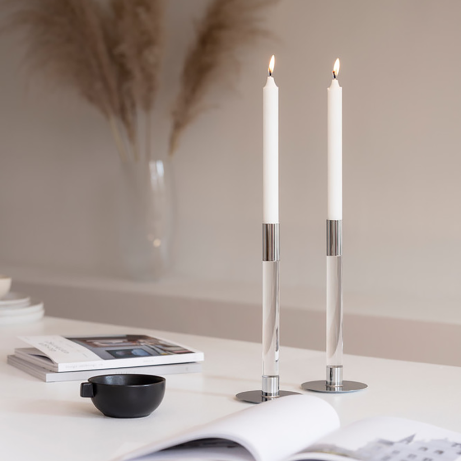 A serene setting features two lit white candles in a set of Orrefors Lumiere Candlestick Silver Medium alongside a black cup, books, and a glass vase with dried plants. Ingegerd Råmans touch adds sophistication.