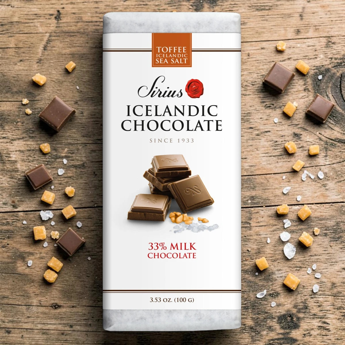 Icelandic chocolate bar labeled “Nói Síríus” on wood, with chocolate squares, toffee pieces, and sea salt flakes scattered around.