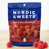 A blue and red Nordic Sweets bag labeled “Soft Raspberries” on a tabletop with Swedish gummy candies shaped like red raspberries displayed in front.