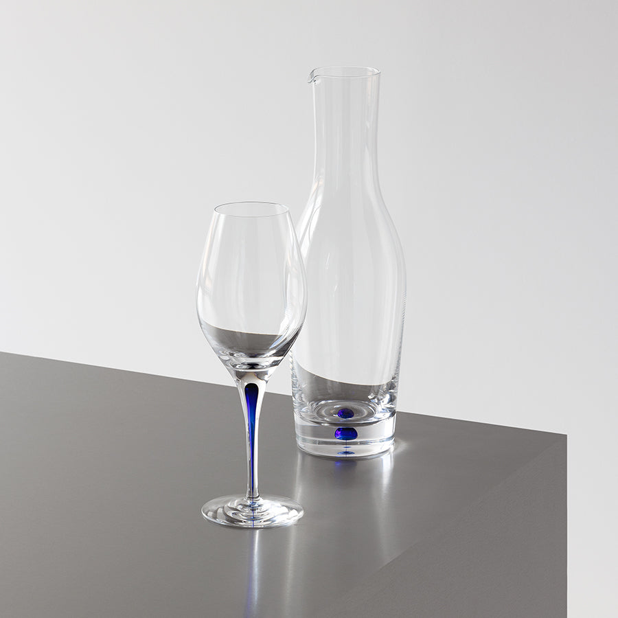 An Orrefors Intermezzo Blue Carafe 37oz by Erika Lagerbielke and a clear wine glass rest elegantly on a gray surface against a white background.