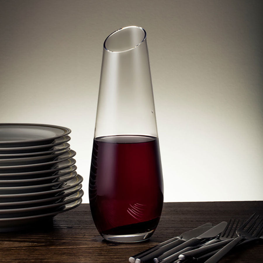 The Orrefors Enjoy Carafe, designed by Erika Lagerbielke and brimming with red wine, sits elegantly on a wooden table beside a stack of plates and cutlery.