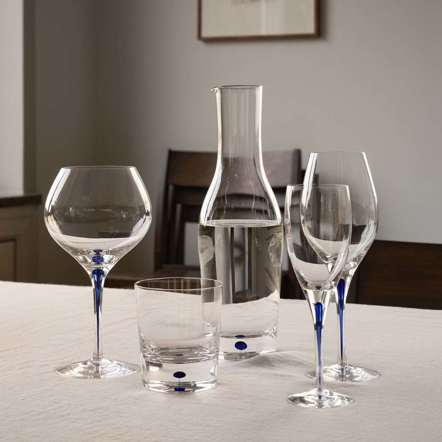 An Orrefors Intermezzo Blue Carafe (37oz) sits elegantly on a table with three matching blue-stemmed wine glasses and a tumbler, contrasting beautifully with the neutral wall and wooden chairs.