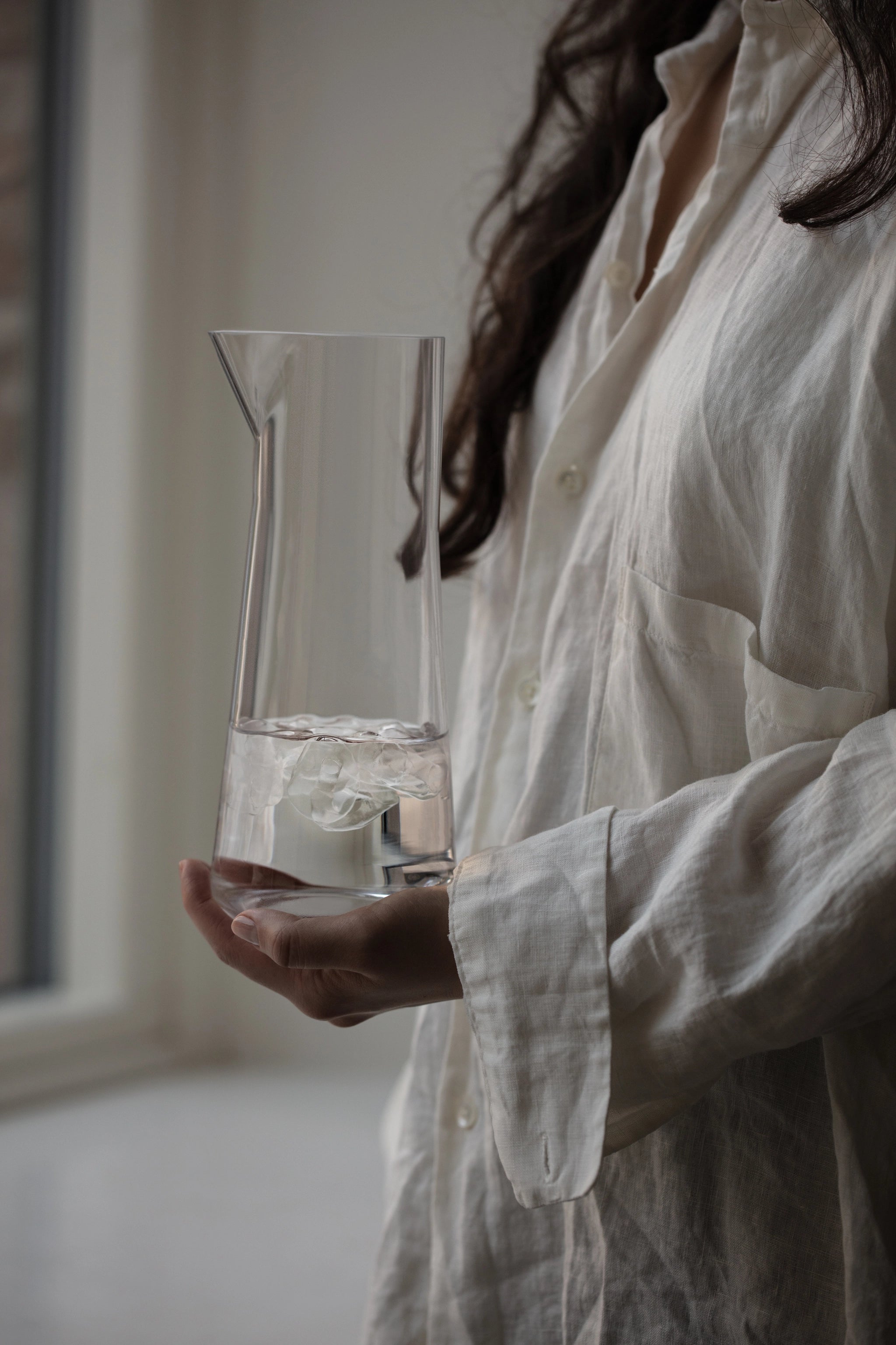 A person in a white shirt holds an Orrefors Informal Clear Carafe with Bowl 34oz containing a flower while standing by a window.