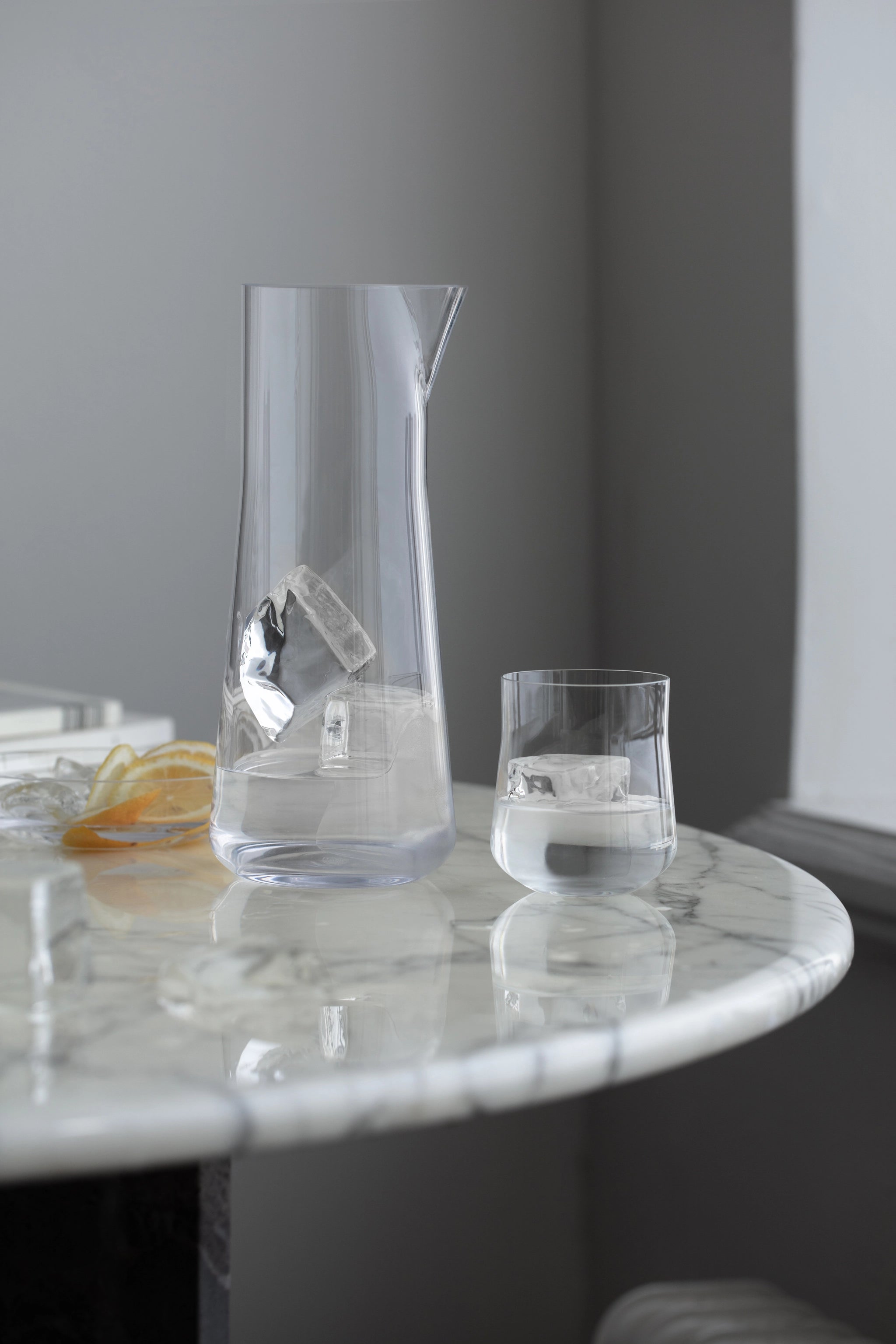 A glass pitcher and glass filled with ice rest on a marble table, surrounded by lemon slices, creating a charming scene reminiscent of an Orrefors display.