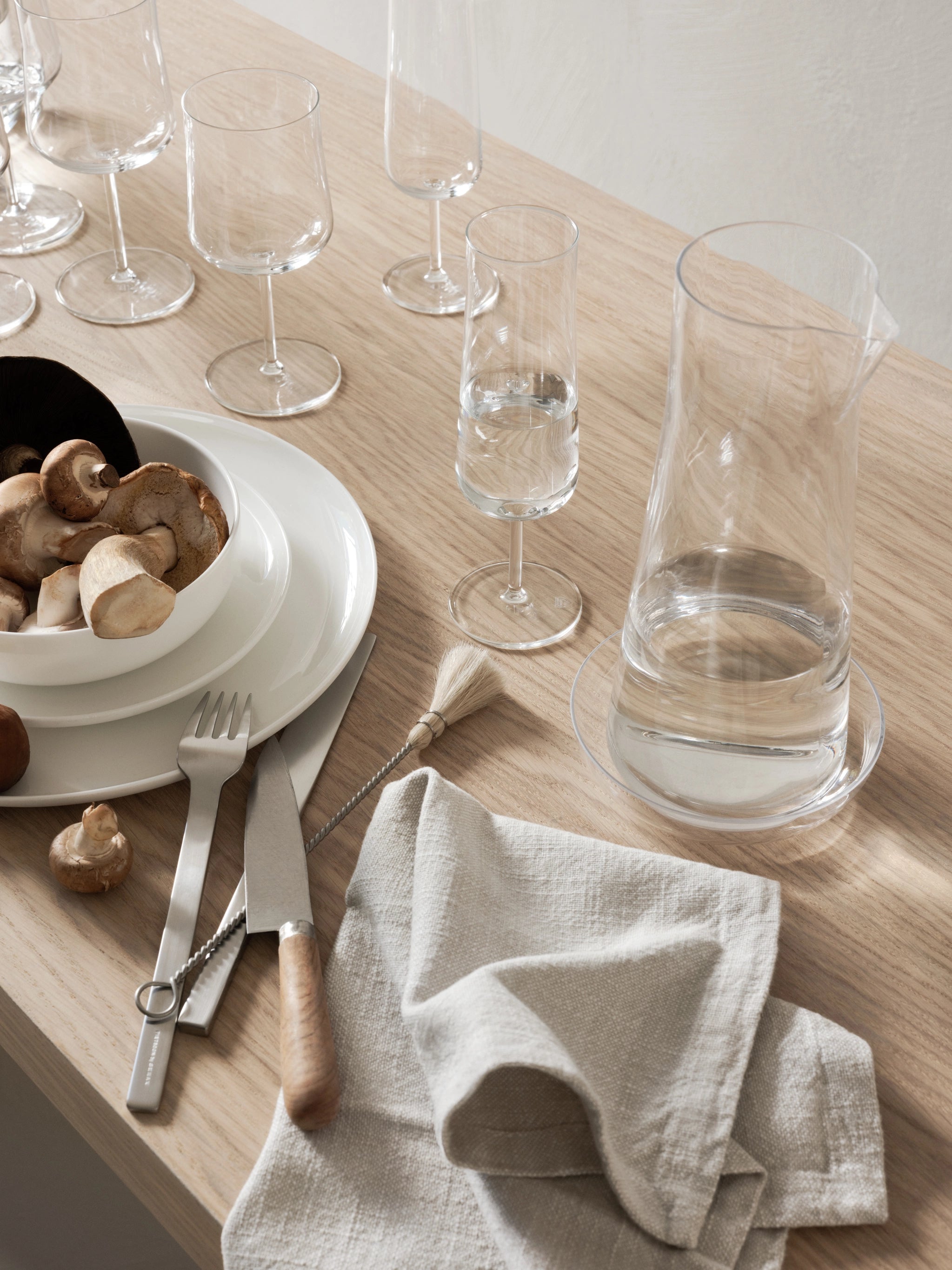 A wooden table set showcases glassware, the Orrefors: Informal Clear Carafe with Bowl 34oz beside a bowl of mushrooms, stacked plates, cutlery, a whisk, and a folded cloth.