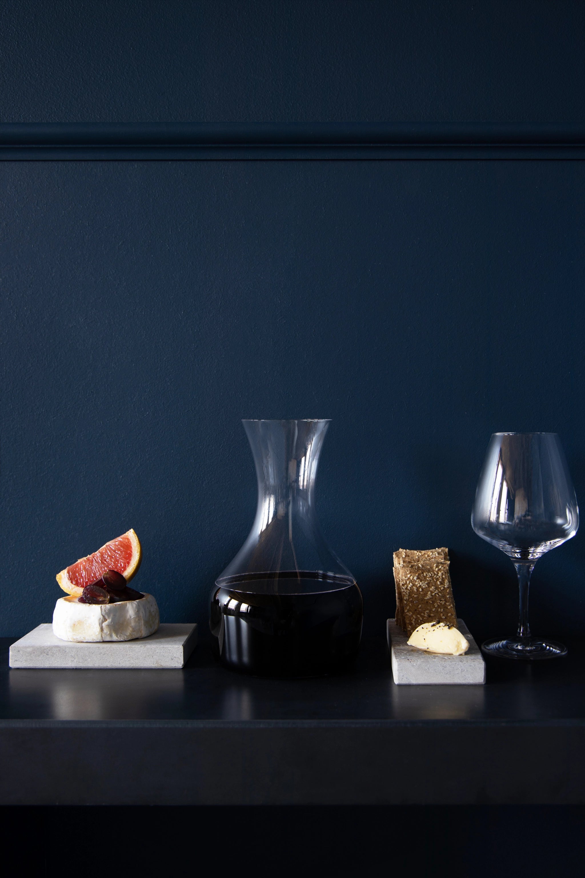 A large 41.5oz Share Carafe by Orrefors and an empty crystal glass sit elegantly on a dark surface, surrounded by crackers with cheese and a cheese wheel decorated with orange slices.