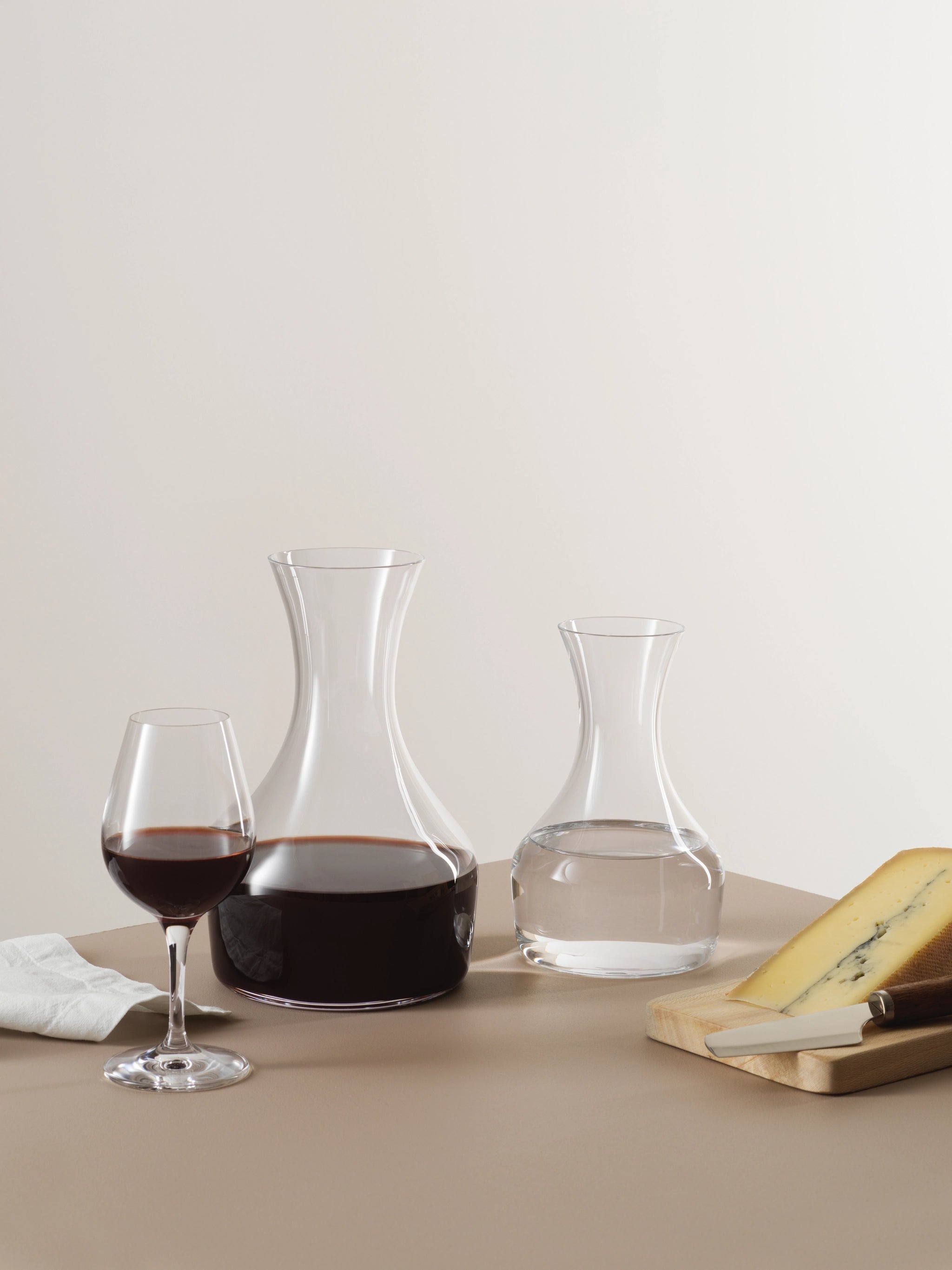 A glass of red wine, an Orrefors Share Carafe with 41.5oz capacity, a crystal water carafe, and a wooden cheese board with sliced cheese and knife rest elegantly on a beige surface alongside a white napkin.