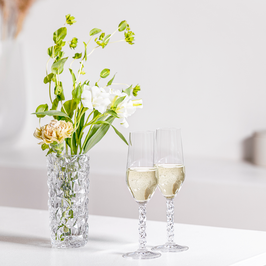 A stunning Orrefors: Carat Vase Small, filled with vibrant flowers, sits elegantly beside two champagne flutes on a pristine white surface.