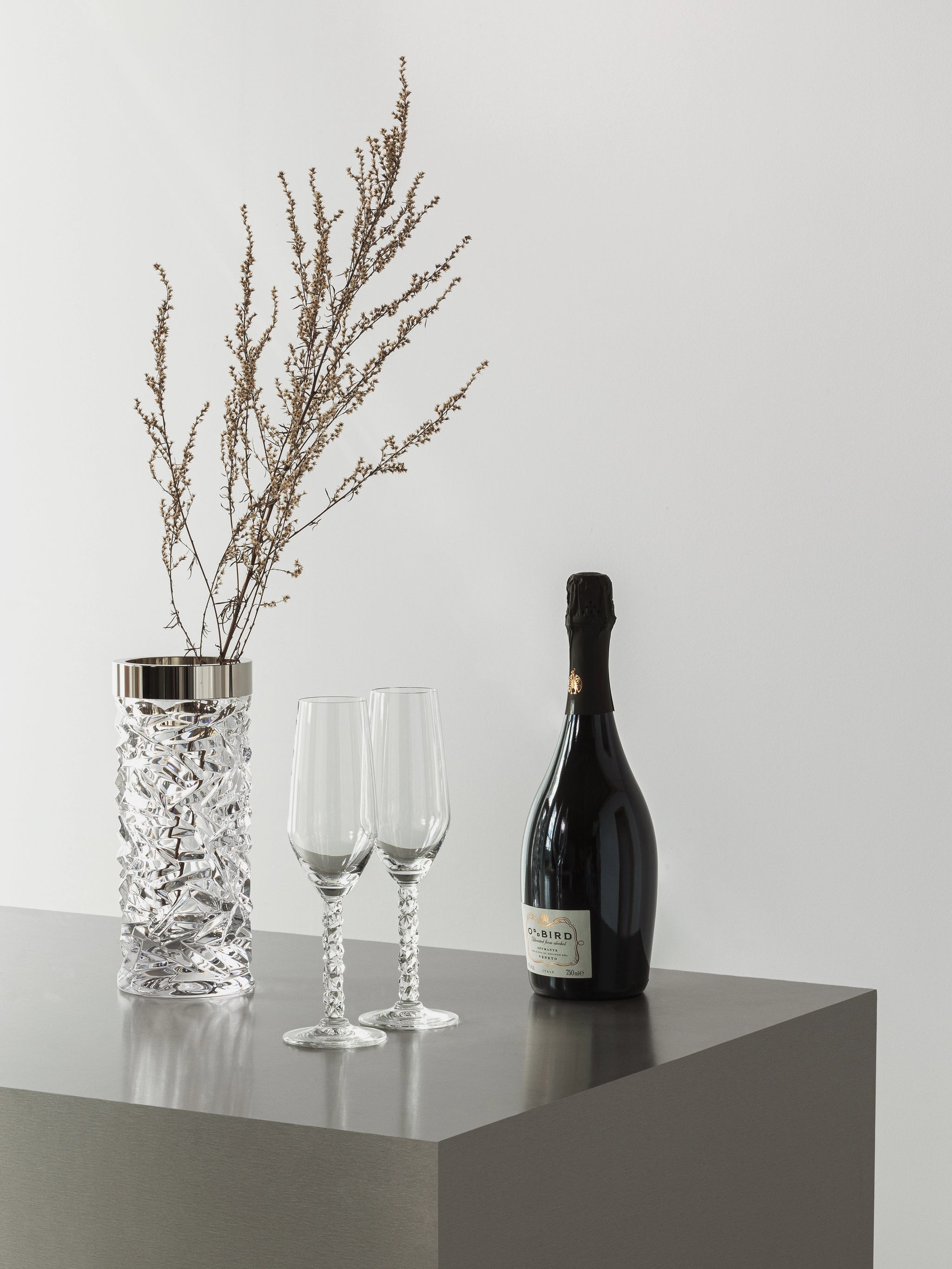 A silver vase with dried branches, Orrefors Carat Champagne Flute 8oz (2-Pack), and an unopened champagne bottle sit elegantly on a gray rectangular table against a white wall.