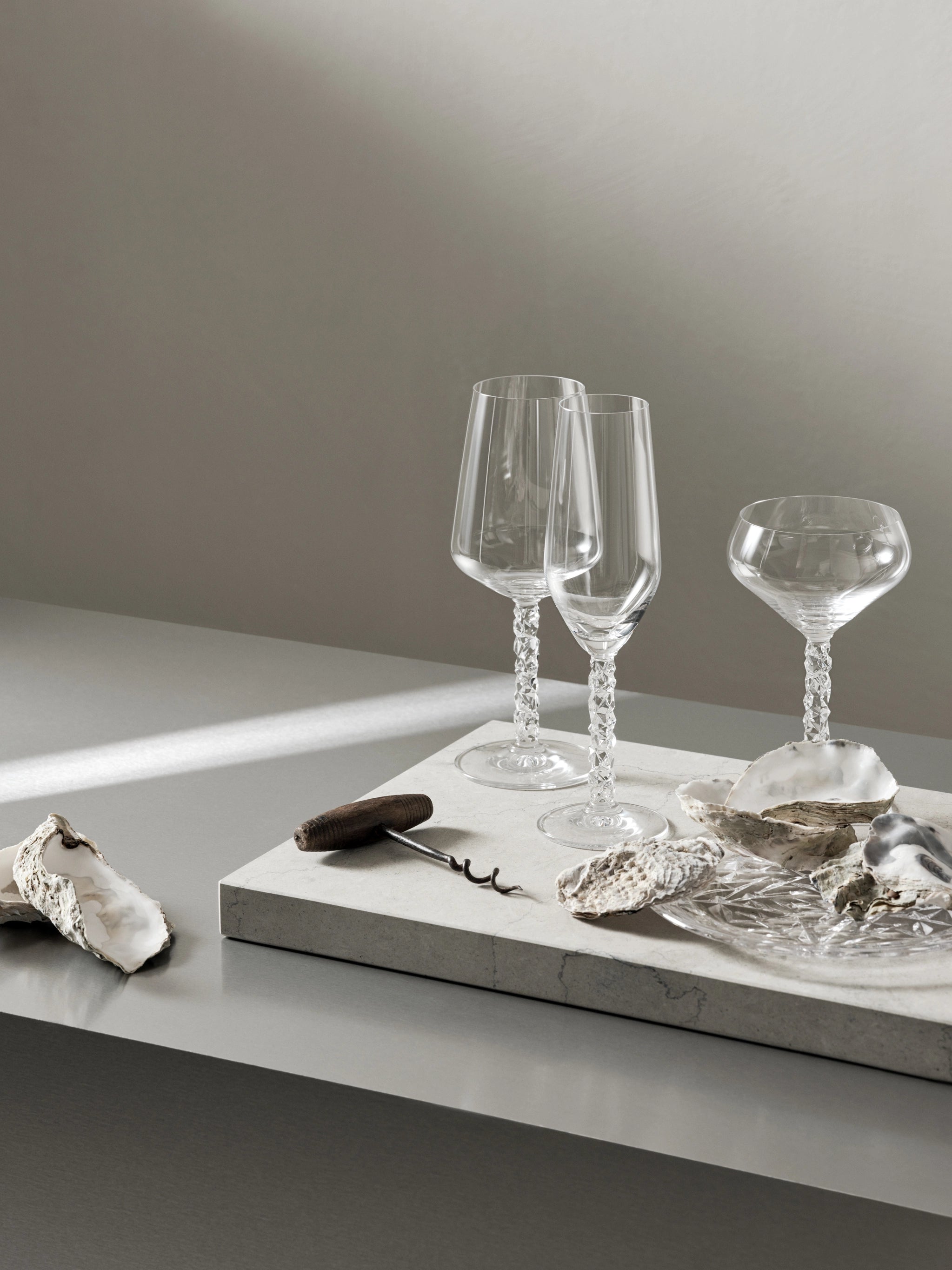 Two elegant Orrefors Carat Champagne Flutes, part of the 8oz (2-Pack) collection, with textured stems, sit gracefully on a stone surface. Surrounded by oyster shells and a mysterious black object, they create a captivating tableau against the minimalist backdrop.