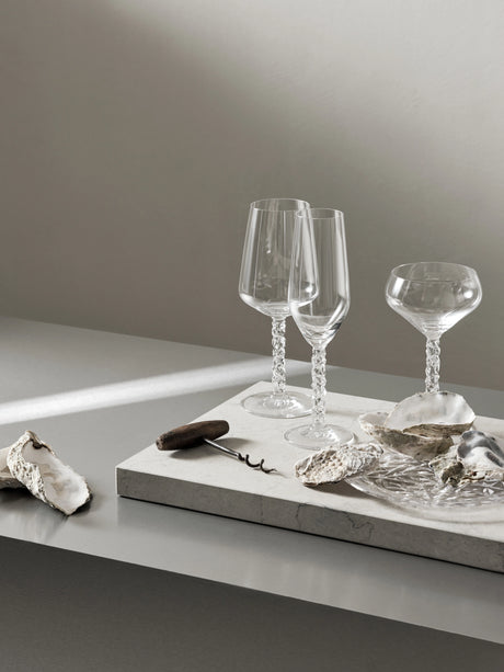 Two elegant Orrefors Carat Champagne Flutes, part of the 8oz (2-Pack) collection, with textured stems, sit gracefully on a stone surface. Surrounded by oyster shells and a mysterious black object, they create a captivating tableau against the minimalist backdrop.