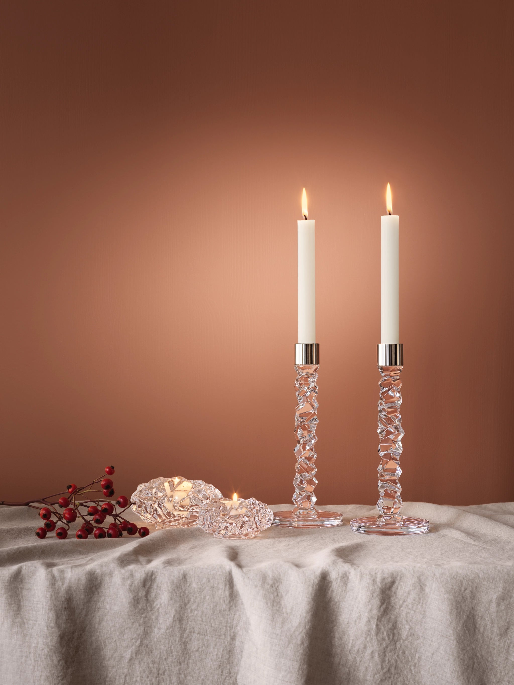 Two lit Orrefors Carat Votive Large Candle Holders adorn a linen-covered surface, accompanied by decorative crystal glass pieces and a sprig of red berries, set against a warm brown background.