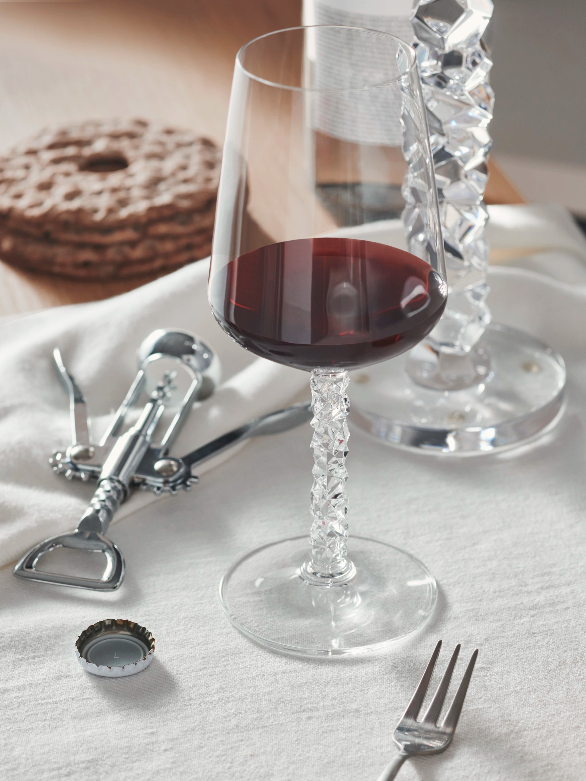 An Orrefors Carat Wine Glass (14.8oz, 2-pack) cradles red wine on a table with a corkscrew, bottle cap, crackers, and fork atop a white cloth.