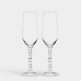 Two elegant Orrefors Carat Champagne Flutes, each 8oz with decorative stems, sit gracefully against a plain white backdrop, epitomizing the sophistication of the Carat Collection.