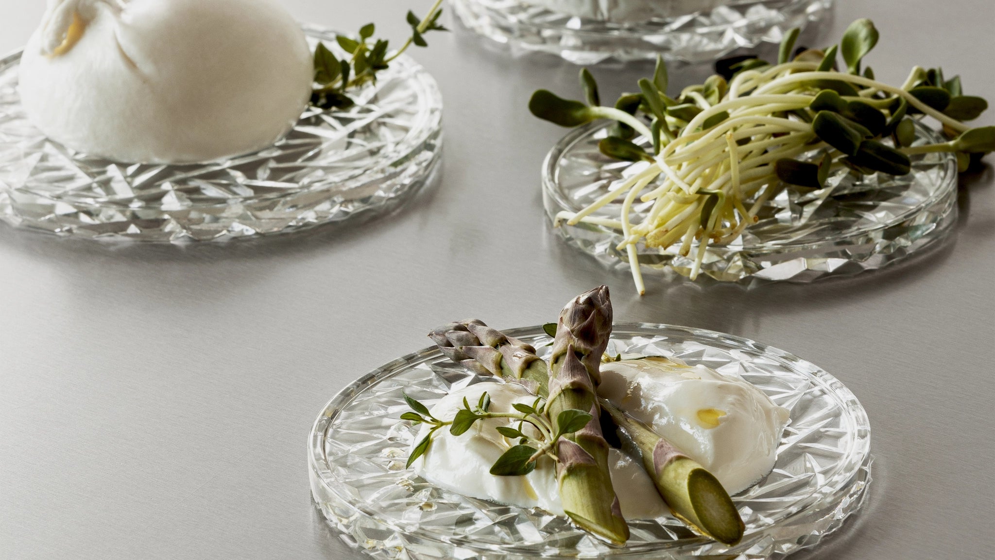 Orrefors Carat Coaster Small (2-Pack) elegantly cradles burrata cheese with asparagus and sprouts on a gray surface.